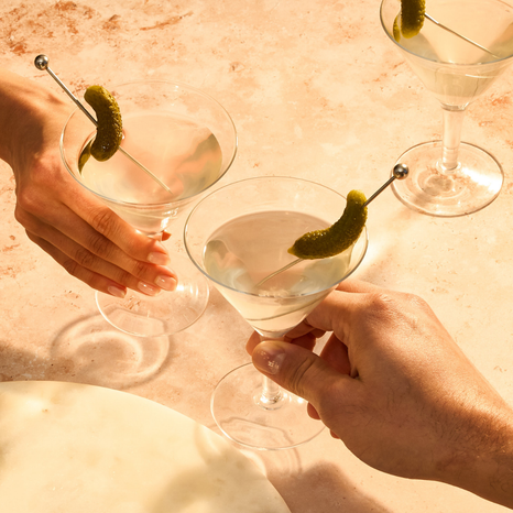 Two hands holding martini glasses with Cornichons picks on a light surface.