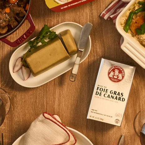 Block of foie gras on a plate with a knife, surrounded by other dishes and a brand label on a wooden table.