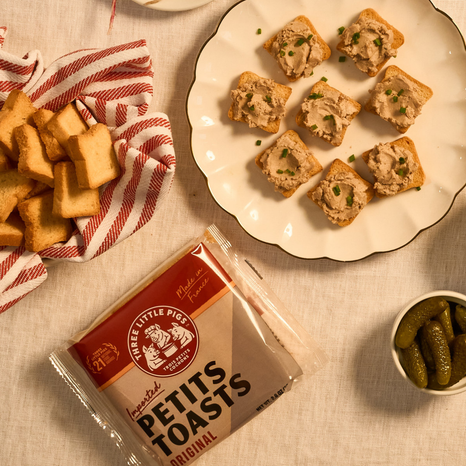 Packaged petits toasts with a plate of toasts and pickles on a table.