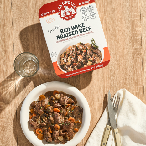 Packaged red wine braised beef on a wooden table with a bowl of beef dish and cutlery.