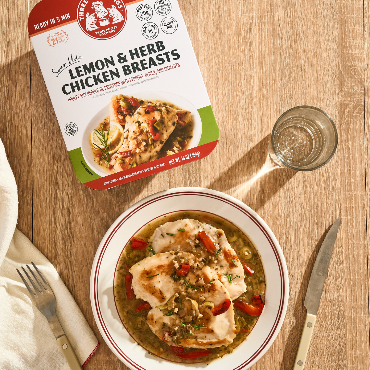 Packaged lemon and herb chicken breasts on a wooden table with a dish of prepared chicken.