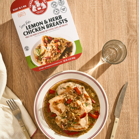 Packaged lemon and herb chicken breasts on a wooden table with a dish of prepared chicken.