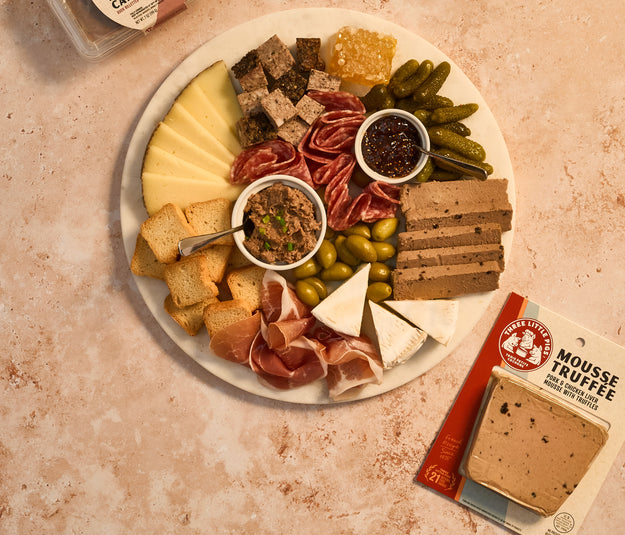 Platter of assorted meats, cheeses, and pickles with a small container of mustard on a textured surface.
