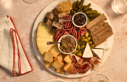 Platter of assorted Charcuterie, cheeses, and Cornichons with people around it on a table.