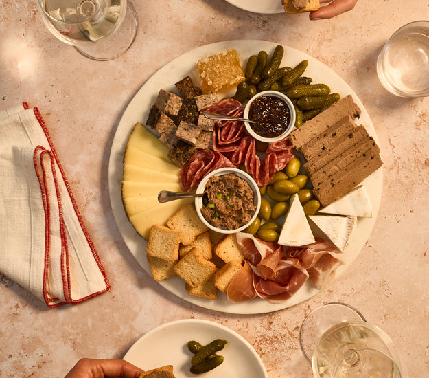 Platter of assorted Charcuterie, cheeses, and Cornichons with people around it on a table.