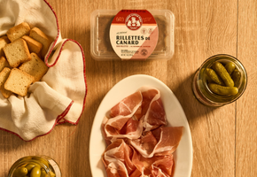 Assorted meats and pickles on a wooden table with a container of Rillettes de Canard.