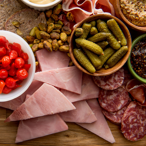 Charcuterie board with varieties of finger foods like slices of Ham, Cornichons, Pepperoni, Nuts and dips.